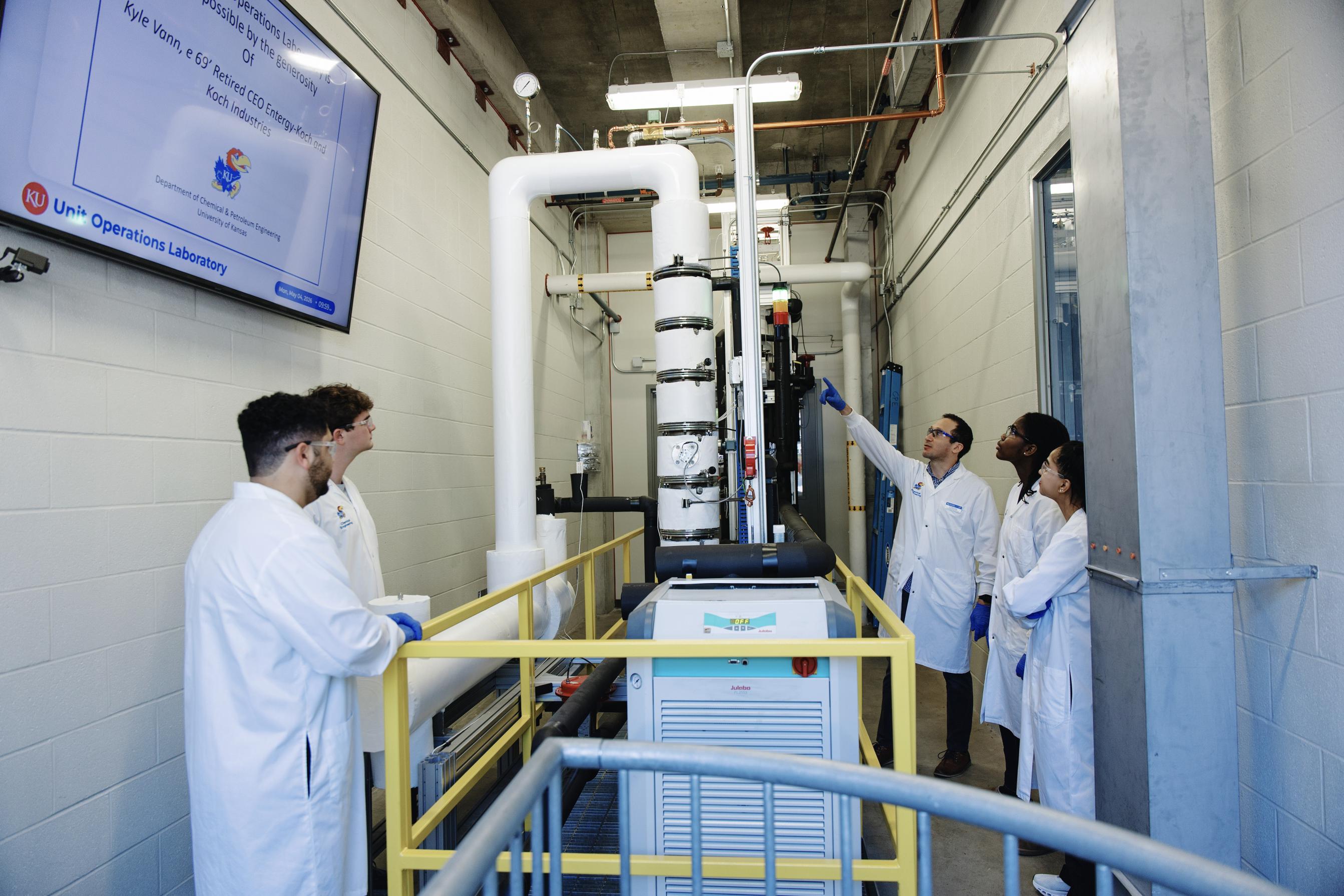 A professor points at parts of the distillation tower for a group of students
