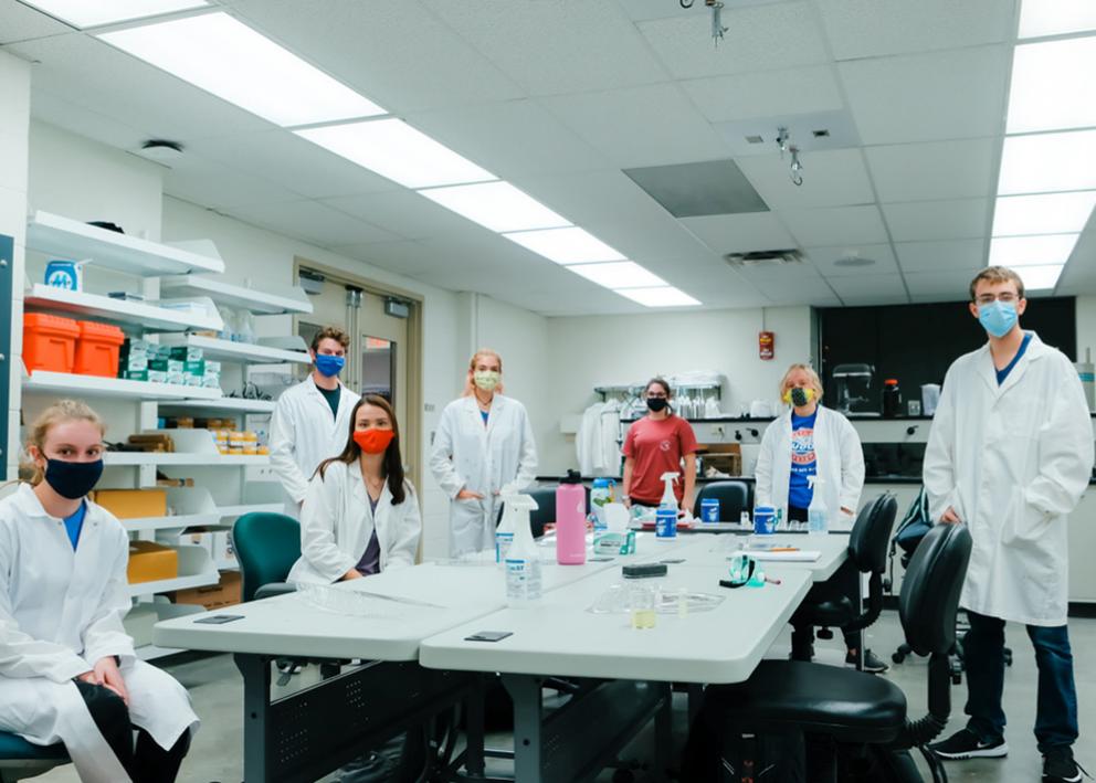 7 students in the lab standing around the lab table wearing masks and social distancing