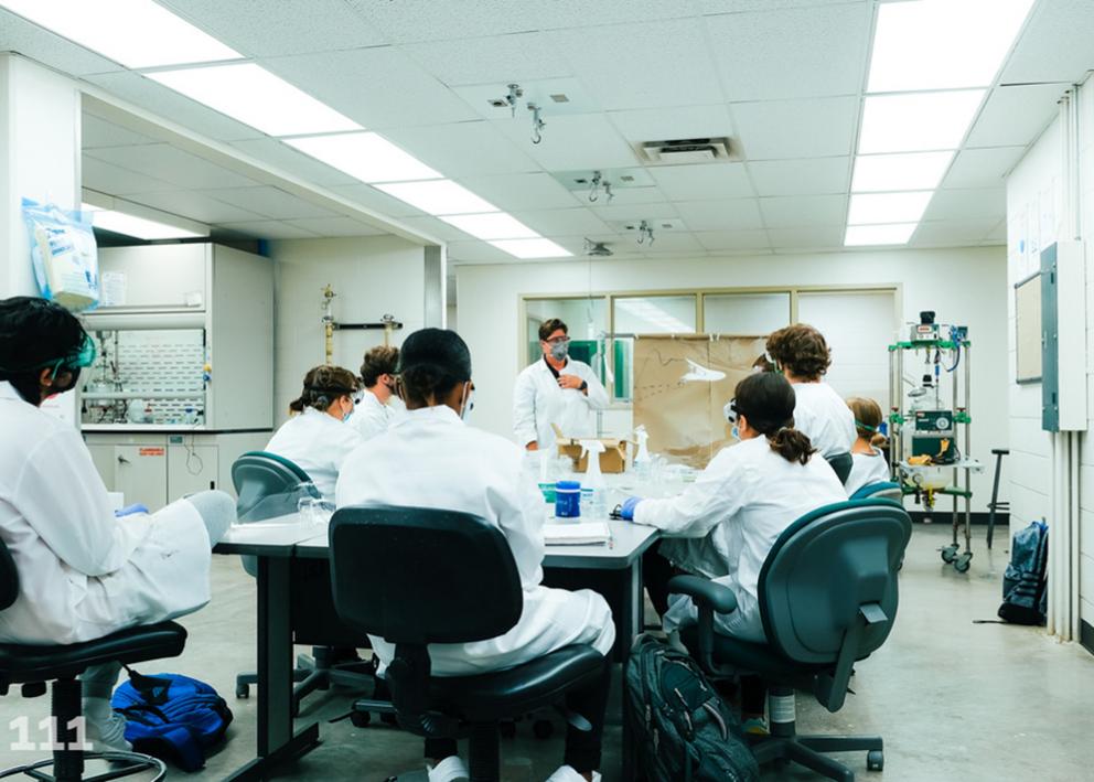 Instructor, Susan Williams, standing in front of a class of eight students sitting in lab coats and lab glasses