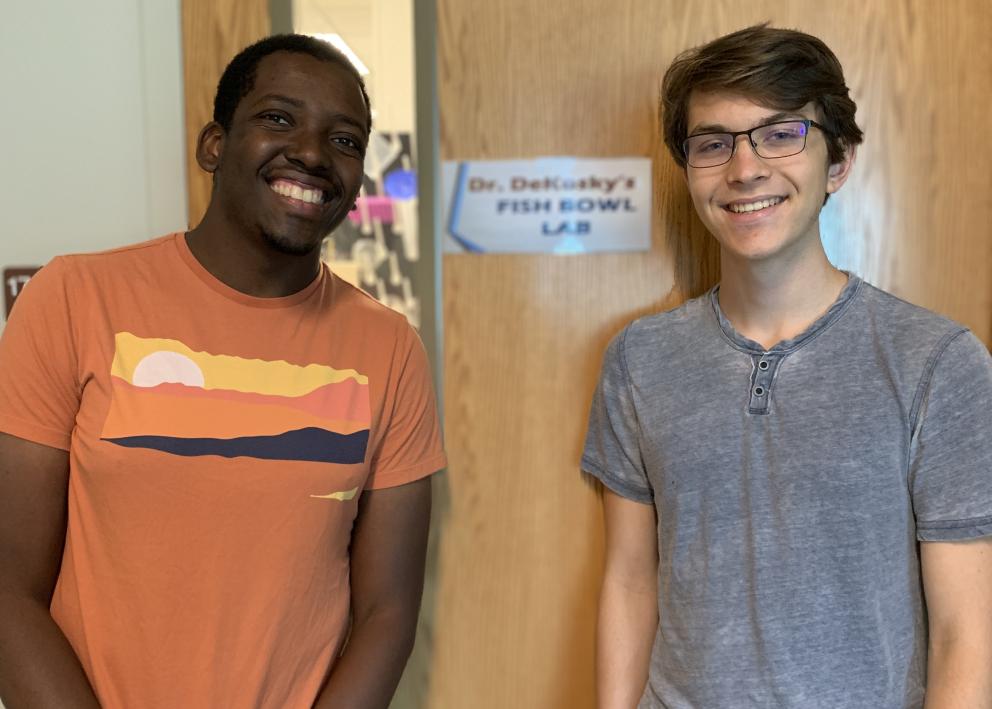 REU student Eli with his graduate student mentor Matheus standing by lab door with sign "Dr. Dekosky's Fish Bowl Lab"