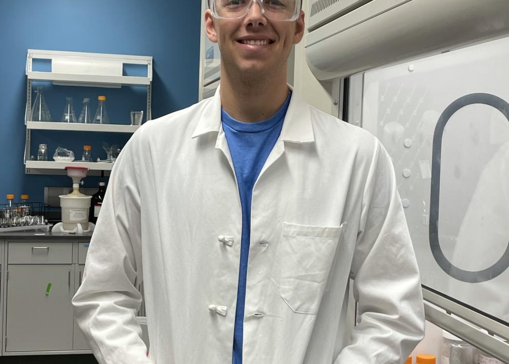 Student in full laboratory safety gear, standing next to fume hood, smiling for picture