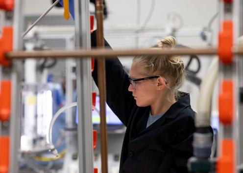 student in the lab holding a device in the air with copper pipes in the foreground