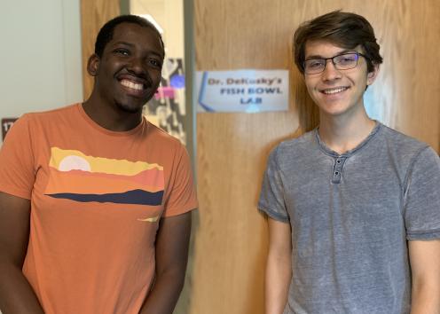 REU student Eli with his graduate student mentor Matheus standing by lab door with sign "Dr. Dekosky's Fish Bowl Lab"