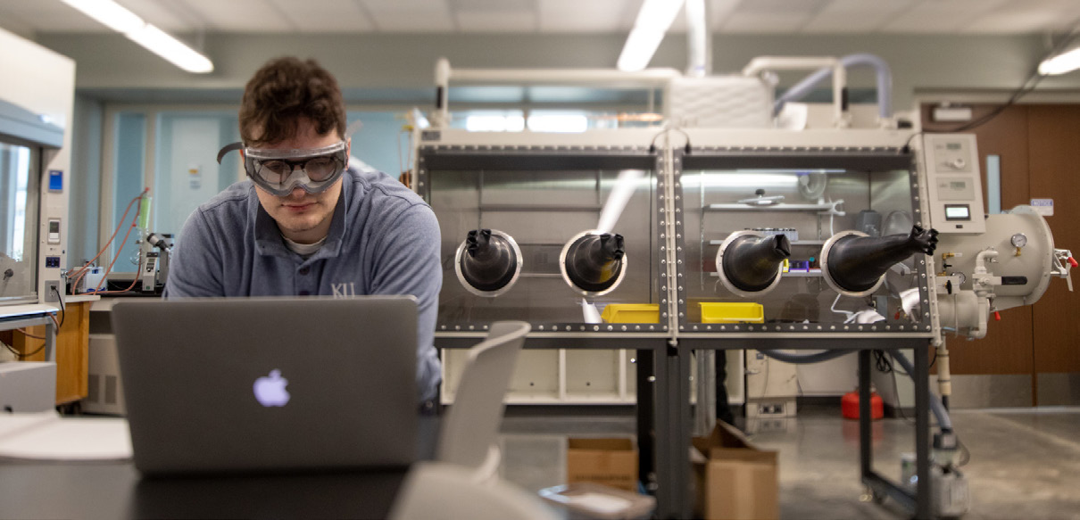 A student works on a laptop with lab equipment in the background